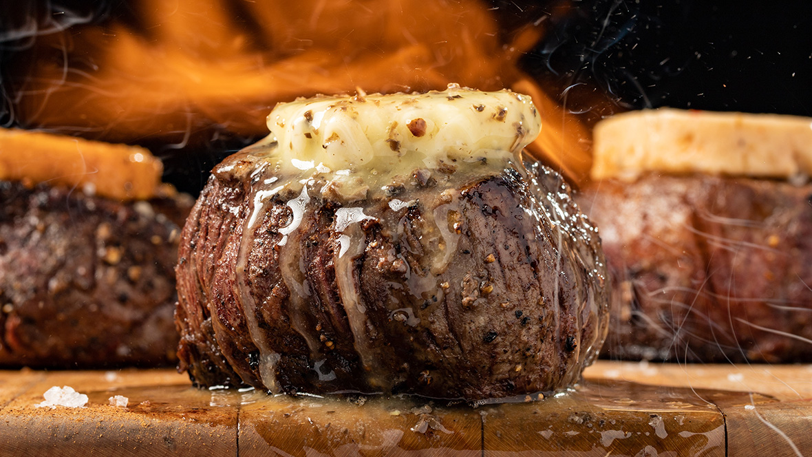 A steak sits grilling on a wood plank in front of a fire. The juicy steak is topped with chopped onions; several other pieces of meat are seen in the background.