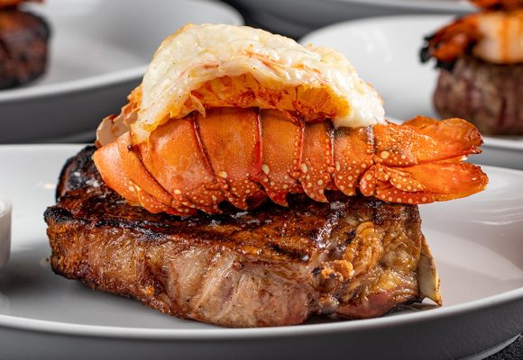 A piece of steak, topped with an orange lobster tail, sits on a clean white plate. To the left of the steak is a sauce in a white dish. Several other plated meals are seen in the background.