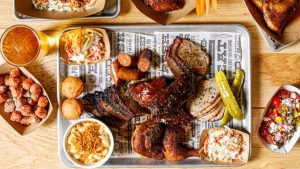The camera is facing down at a wooden dinner table, and several dishes and beverages from The Polite Pig are scattered around the image. Some dishes are in cardboard boats, while others presented on a silver tray covered in faux newspaper. Food depicted include sliced meats, chicken, homemade macaroni & cheese topped with breadcrumbs, and more.