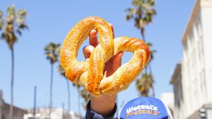 A person holds up an oversized, salted Wetzel&rsquo;s Pretzel above their head; you can see just the top of their blue Wetzel&rsquo;s Pretzels hat. The background is a blue sky with palm trees and buildings lining the sides of the street.