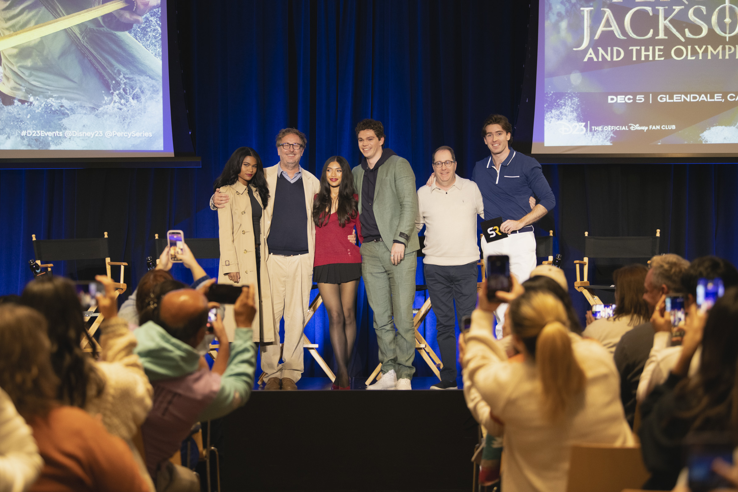 A photo of panelists from D23&rsquo;s Percy Jackson and the Olympians Spotlight Series event in Glendale, CA. From left to right are Tamara Smart, Craig Silverstein, Dior Goodjohn, Daniel Diemer, James Bobin, and Liam Crowley. Fans below clamor to take photos, and talent pose onstage in front of a row of black director&rsquo;s chairs and large projections of the event logo.