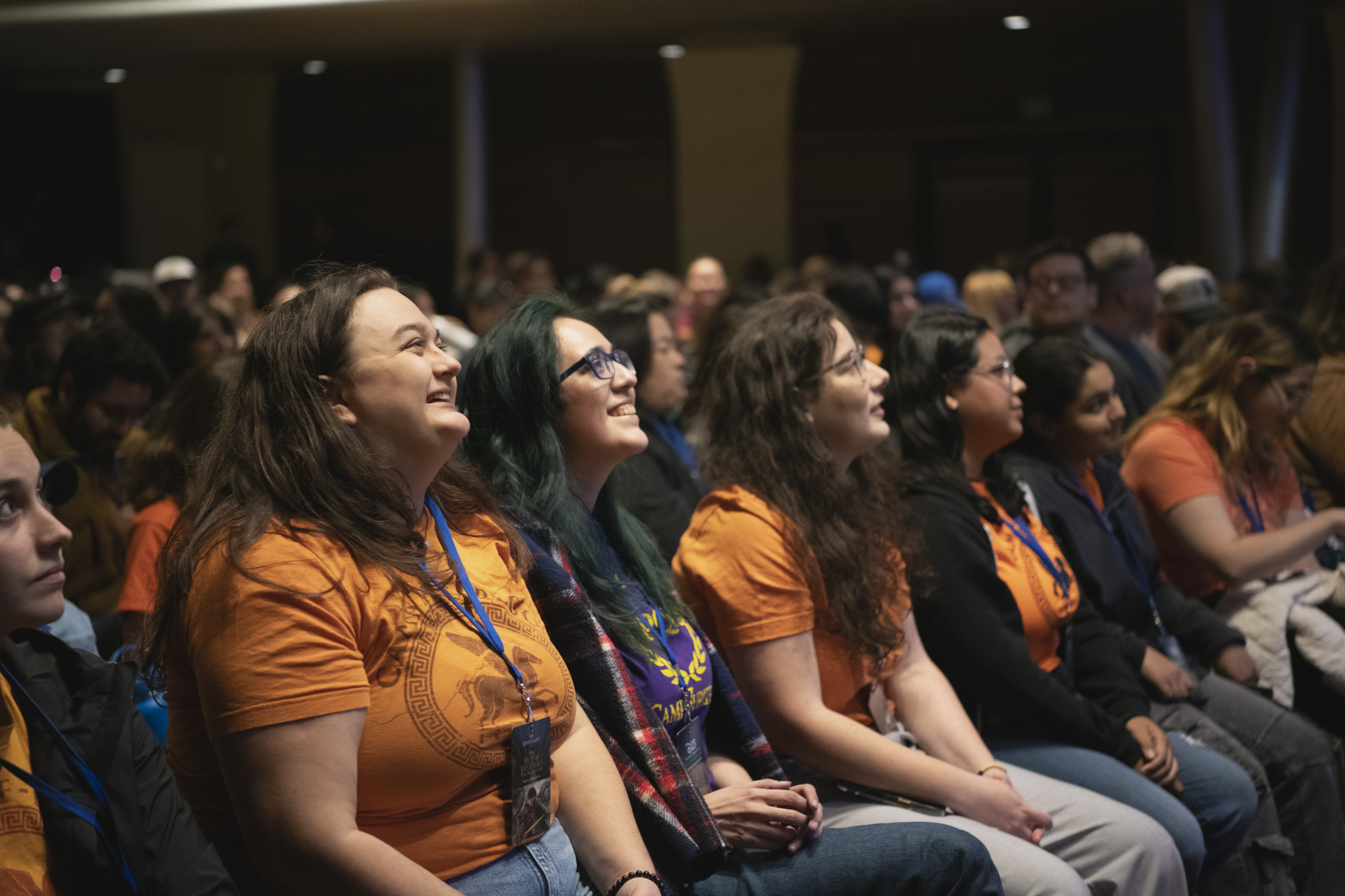 A close-up of audience members at D23&rsquo;s Percy Jackson and the Olympians Spotlight Series event in Glendale, CA. In a row, young women in orange Camp Half-Blood t-shirts smile and attentively watch onstage pre-show content. There is an air of excitement and anticipation.