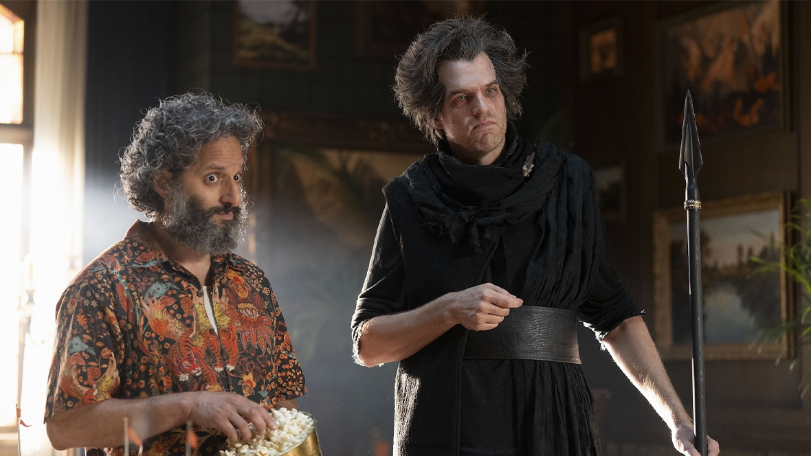 Mr. D, played by Jason Mantzoukas, eats popcorn out of a golden bucket while Tantalus, played by Timothy Simons, holds a spear in Percy Jackson and the Olympians.