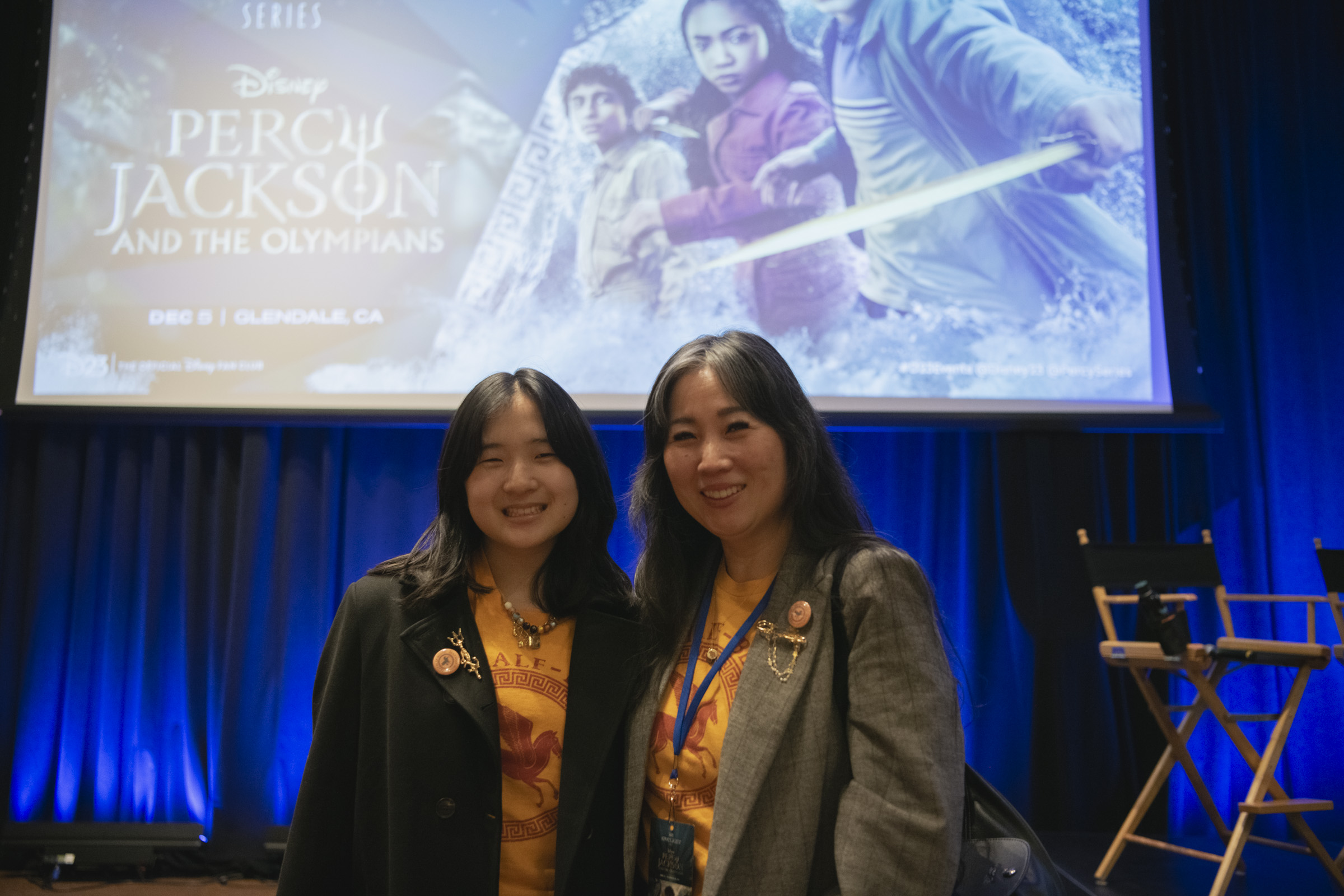 A duo clad in Camp Half-Blood gear at D23&rsquo;s Percy Jackson and the Olympians Spotlight Series event in Glendale, CA. The background includes a large projected image of the event logo, featuring the series&rsquo; main trio. Both guests wear blazers decorated with buttons and small gold accessories evoking ancient Greek jewelry.