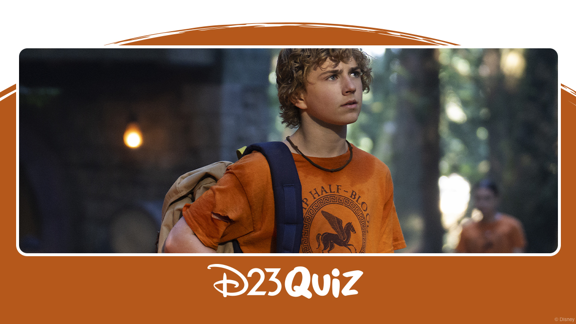 Percy Jackson stands in front of a cabin at Camp Half-Blood, wearing his backpack on one shoulder with a contemplative expression on his face. He is wearing an orange Camp Half-Blood T-shirt, as are the out-of-focus campers in the background of the image.