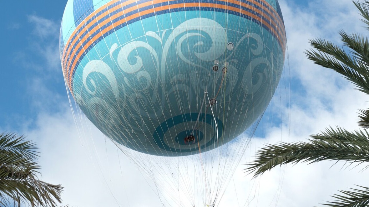 A massive turquoise hot air balloon is being lifted into the blue Orlando sky. The balloon is surrounded by a net and has accents of dark blue and orange stripes. Around the image of the balloon, on either side and in the foreground, are palm tree fronds.