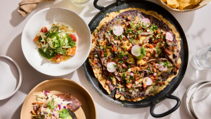 Traditional Mexican dishes are displayed on a table, covered with a white linen tablecloth. A tlayuda with steak takes up a majority of the table. Other dishes include tacos and a basket of chips, seen just at the upper right edge of the photo.