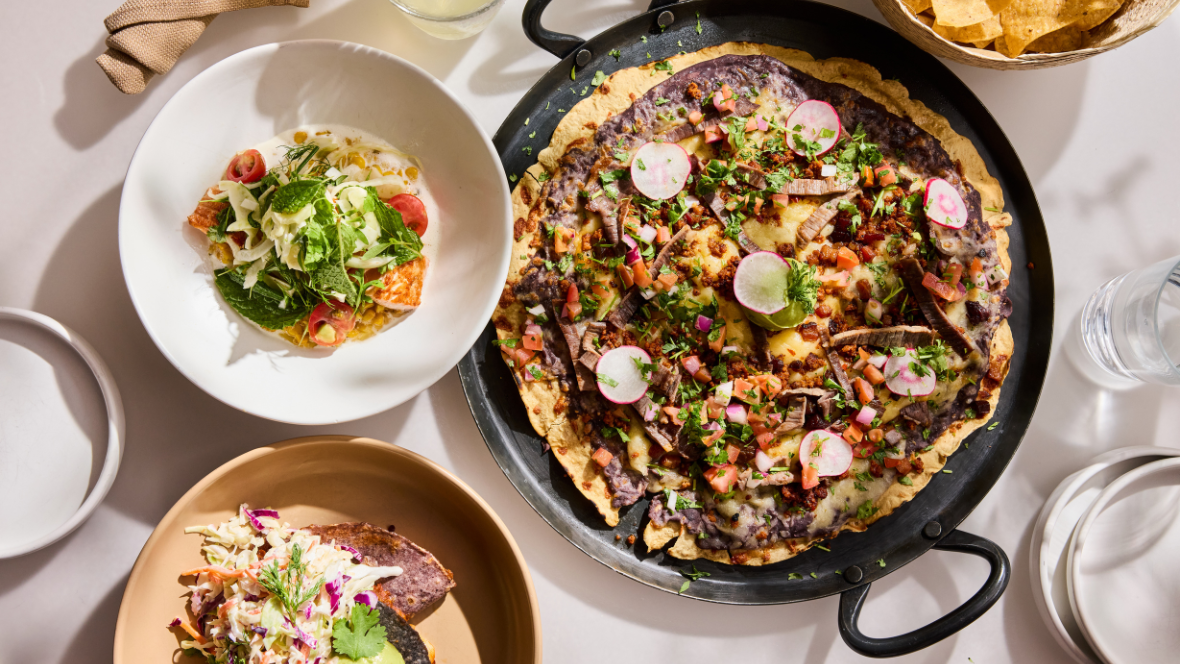 Traditional Mexican dishes are displayed on a table, covered with a white linen tablecloth. A tlayuda with steak takes up a majority of the table. Other dishes include tacos and a basket of chips, seen just at the upper right edge of the photo.