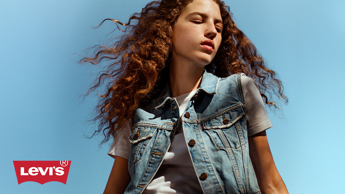 A young woman with wavy hair is looking down in front of a blue background. She is wearing a white shirt and a light blue Levi&rsquo;s jean vest.