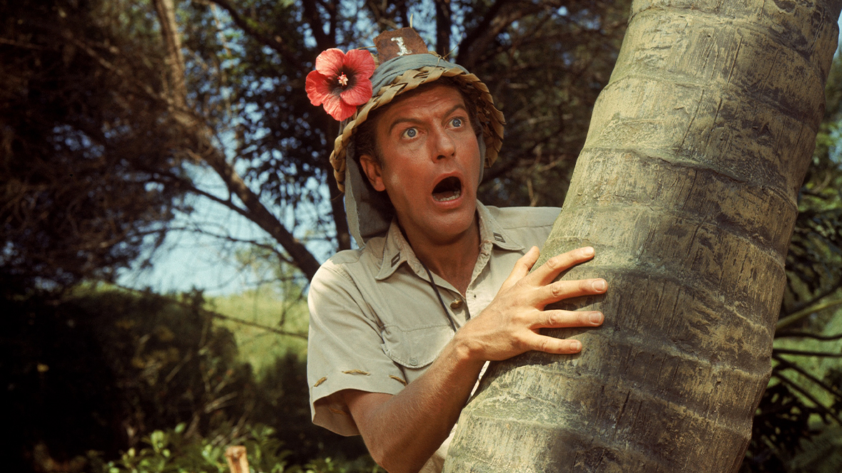 An image of Disney Legend Dick Van Dyke from 1966’s Lt. Robin Crusoe U.S.N. Van Dyke is wearing a wicker hat with a flower and making a funny face, looking at something off camera.