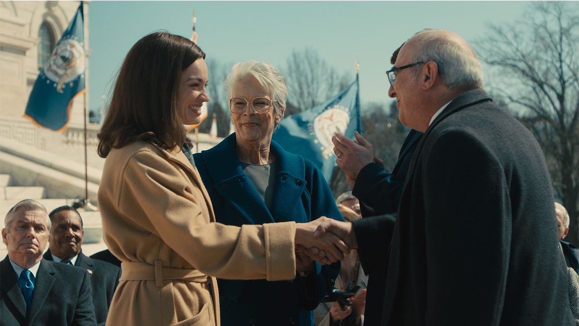 In an image from 20th Century Studios’ Ella McCay, Ella (Emma Mackey), left, is shaking the hand of an older man to her right. Between her and the older man stands her aunt Helen (Disney Legend Jamie Lee Curtis), who is smiling at Ella with pride. The group is standing outside of a state house; flags can be seen flying in front of the state house’s steps, and a crowd of people is looking on at the scene behind Ella and Helen.