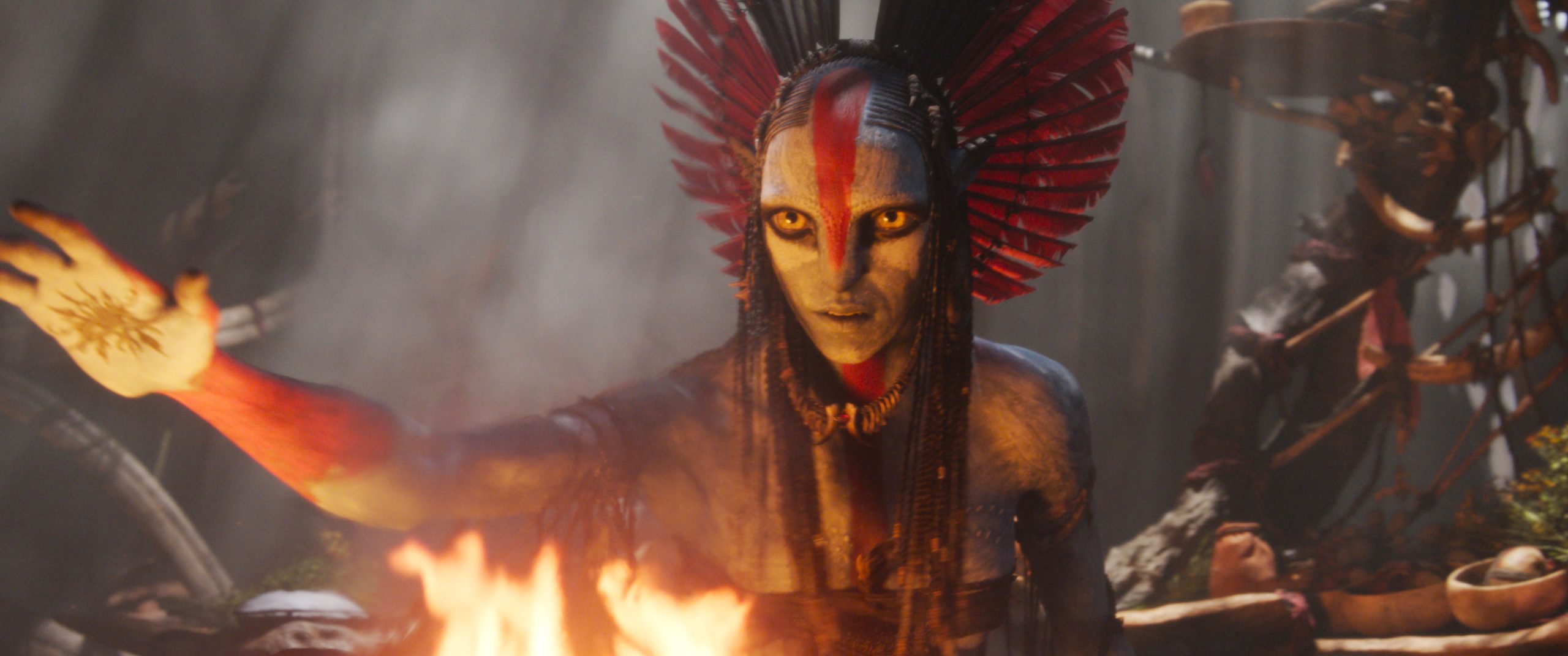 In an image from 20th Century Studios&rsquo; Avatar: Fire and Ash, Varang (Oona Chaplin) is speaking to someone off camera to the left. One of her hands is raised; you can see a tattoo on that palm. Flames of a fire can be seen in front of her, and she has an intense look on her face.