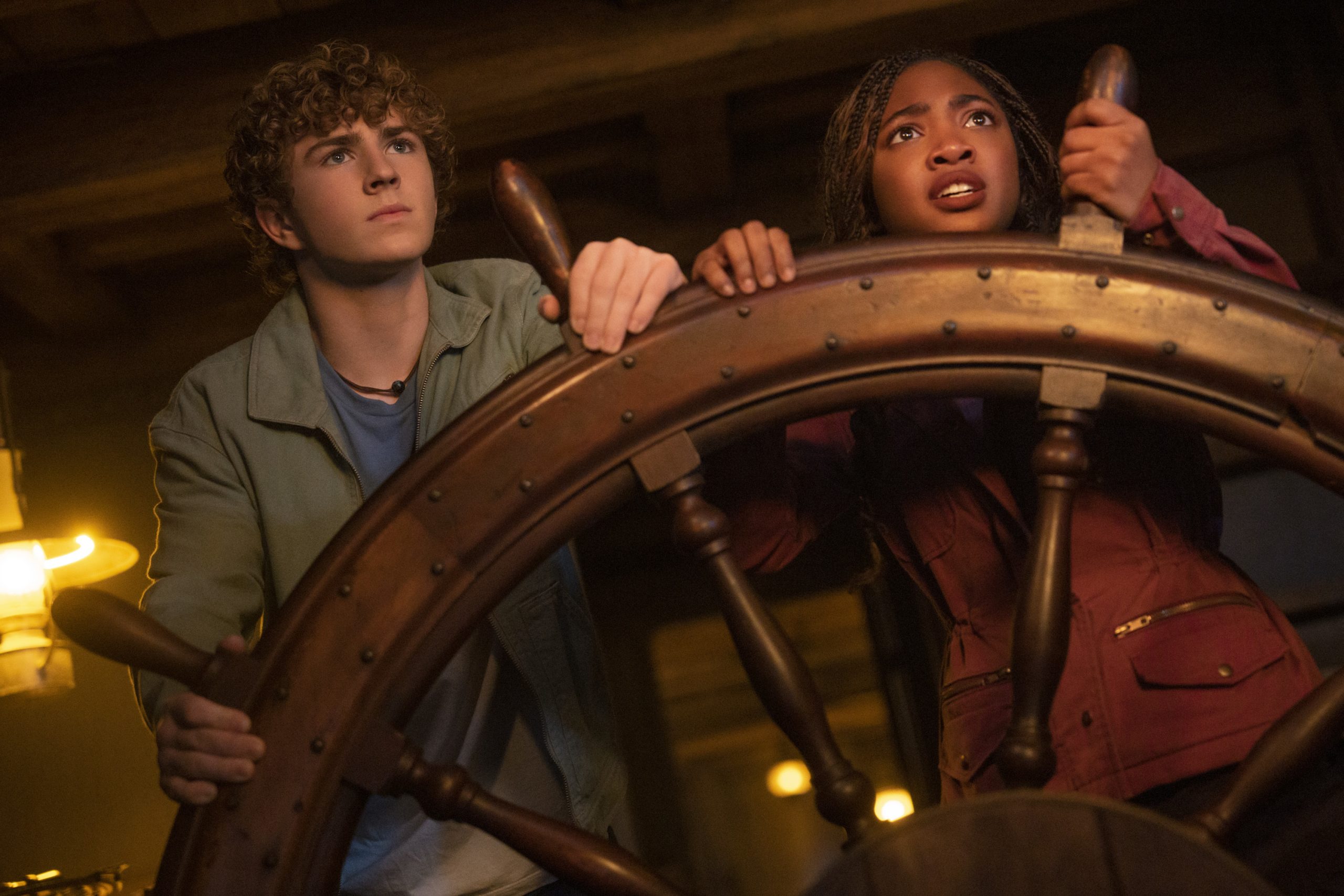 In an image from Season 2 of Disney+’s Percy Jackson and the Olympians, Percy (Walker Scobell) and Annabeth (Leah Sava Jeffries) are seen behind a large ship’s wheel. They are looking intently at something off screen to the right.