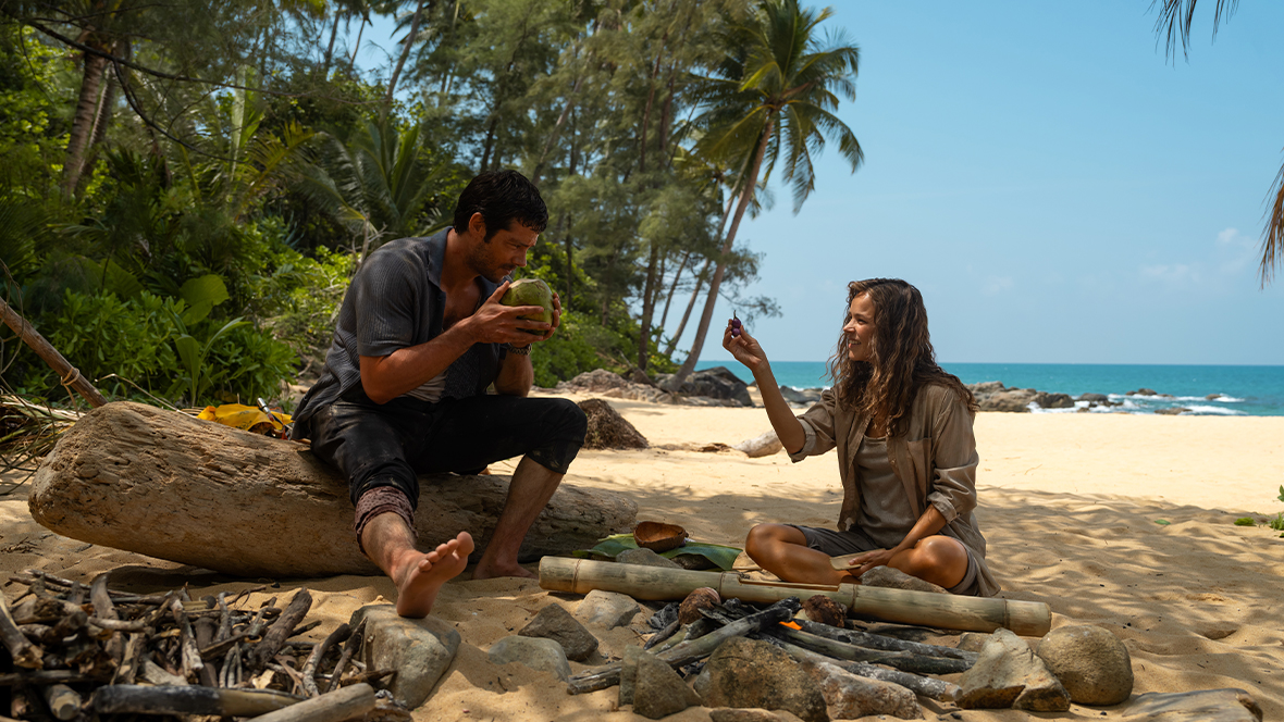 Bradley Preston, played by Dylan O'Brien, drinks from a coconut on a deserted beach as Linda Liddle (Rachel McAdams) holds berries in 20th Century Studios' Send Help.