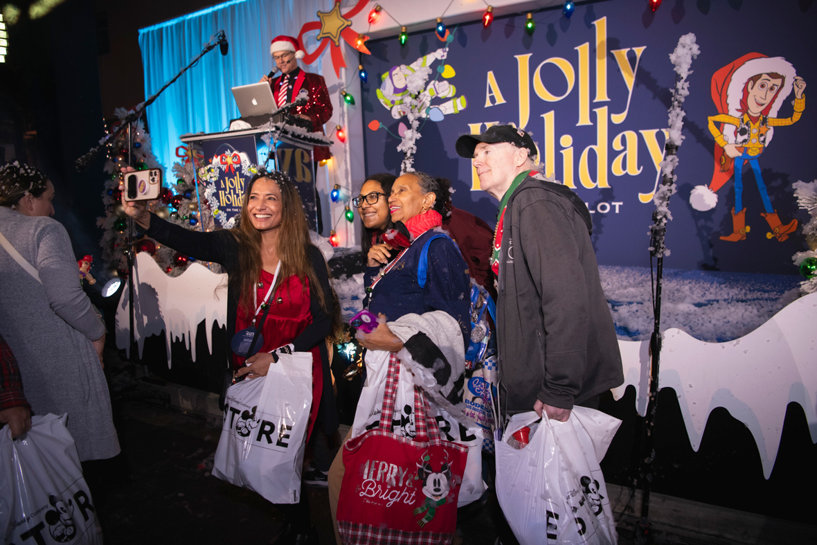 A group of attendees of A Jolly Holiday with D23 in Burbank, CA, take a selfie in front of the main stage. They wear jackets and hold shopping bags with The Walt Disney Company Store logo. The stage is decorated with cutouts evoking snowfall, and string lights around an event logo featuring Buzz Lightyear and Woody.