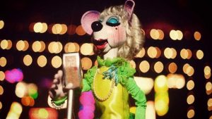 In an image from Bob Baker Marionette Theater, a mouse marionette dressed in yellow and green sings into a microphone. Stage lights glow behind her.