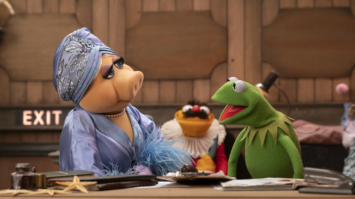 A still from the brand-new special event The Muppet Show, airing February 4, 2026, on Disney+. Miss Piggy, left, is wearing a blue turban and robe, and is talking to Kermit, right. In between and behind them, Lew Zealand can be seen watching the conversation.