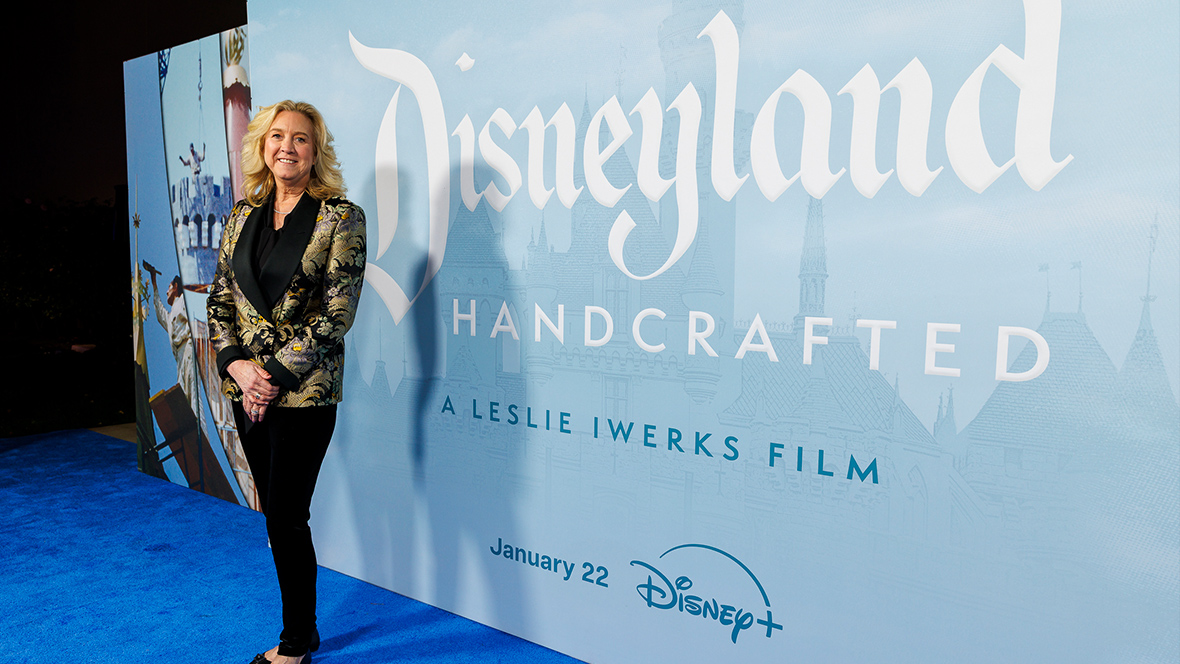 Director Leslie Iwerks stands on the blue carpet at the premiere of her documentary film Disneyland Handcrafted. A step and repeat backdrop, featuring the film&rsquo;s logo, is behind her and to the right. She smiles at the camera.