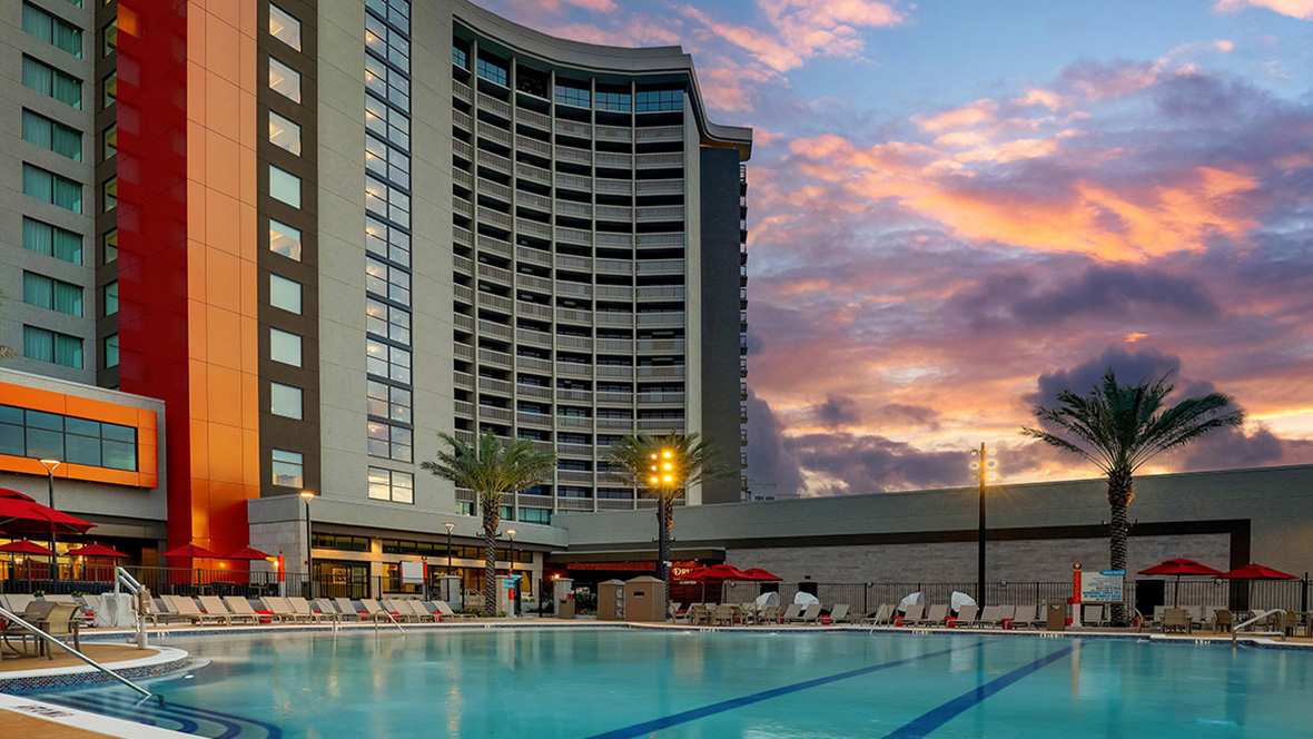 Resort-style outdoor swimming pool at Drury Plaza Hotel Orlando &ndash; Disney Springs Area, with lounge chairs, palm trees, and the hotel building exterior at sunset.