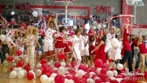 From left to right: Sharpay, Chad, Troy, Gabriella, Taylor, and Ryan during &ldquo;We&rsquo;re All in This Together,&rdquo; the final number of High School Musical (2006). Sharpay is wearing a white tank top and white jeans. Chad is wearing a white East High basketball uniform. Troy is wearing a white East High basketball track suit. Gabriella is wearing a red dress that falls just above the knee. Taylor is wearing a white button-up with a red and white striped tie and a white pleated skirt. Ryan is wearing a red fedora, red button up, and black pants. Red and white ballons surround their feet and confetti fills the air. Behind them, the rest of the student body of East High is cheering.