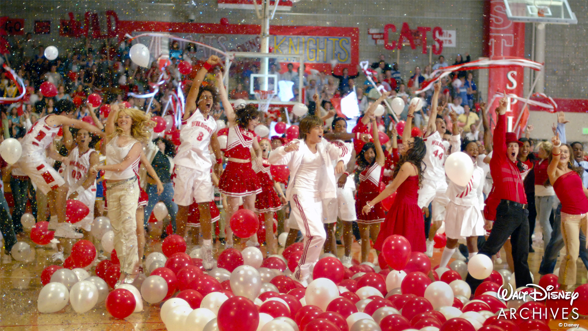 From left to right: Sharpay, Chad, Troy, Gabriella, Taylor, and Ryan during &ldquo;We&rsquo;re All in This Together,&rdquo; the final number of High School Musical (2006). Sharpay is wearing a white tank top and white jeans. Chad is wearing a white East High basketball uniform. Troy is wearing a white East High basketball track suit. Gabriella is wearing a red dress that falls just above the knee. Taylor is wearing a white button-up with a red and white striped tie and a white pleated skirt. Ryan is wearing a red fedora, red button up, and black pants. Red and white ballons surround their feet and confetti fills the air. Behind them, the rest of the student body of East High is cheering.