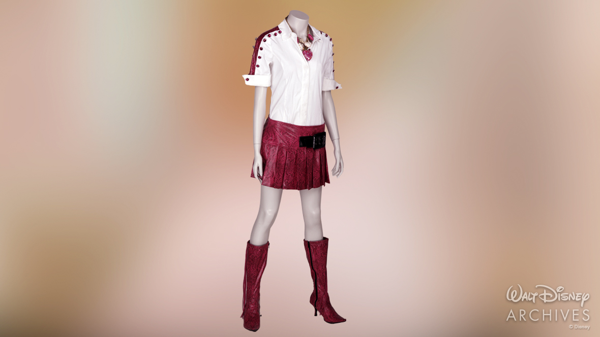 Sharpay&rsquo;s outfit is displayed on a mannequin. Her shirt is a short-sleeve white button-up with faux pink snakeskin along the top of the shoulders. Along the edge of the snakeskin are large pink rhinestone buttons. Her pleated skirt is made of the same pink snakeskin and is mid-thigh length. A chunky black accent belt sits on the left side of the skirt. Her knee-high stiletto boots are made of the same pink snakeskin and feature long pink tassels on the side. Her necklace features alternating pink and white chunky bedazzled hearts and a gold chain. 