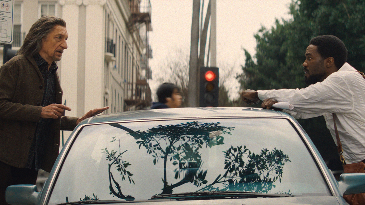In a still from Marvel Television&rsquo;s Wonder Man, Trevor Slattery (Sir Ben Kingsley), left, and Simon Williams (Yahya Abdul-Mateen II), right, are leaning on opposite sides of a parked car and talking. It is overcast, and a stoplight can be seen behind and between them.