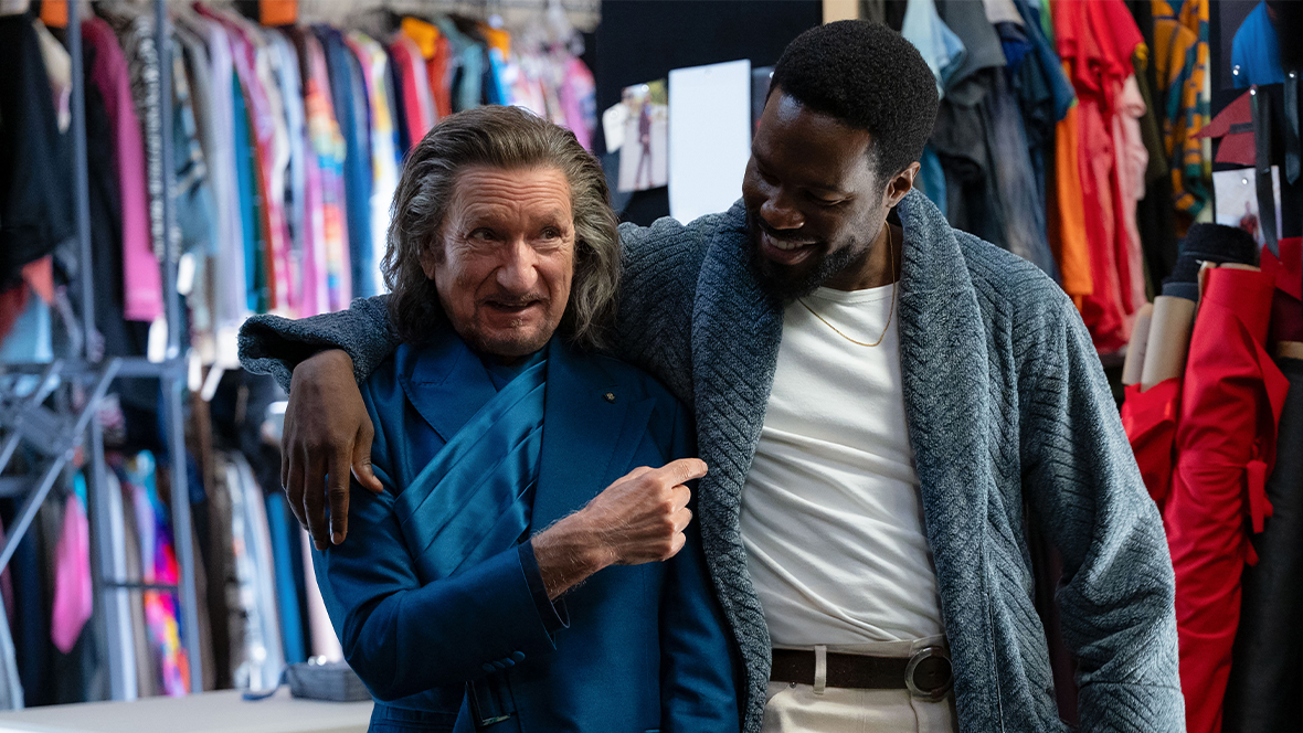 In a still from Marvel Television’s Wonder Man, Trevor Slattery (Sir Ben Kingsley), left, and Simon Williams (Yahya Abdul-Mateen II), right, are in a film studio’s costume department; Simon has a grey sweater on that Trevor—who is slightly behind Simon—is holding onto the lapel of and smiling.