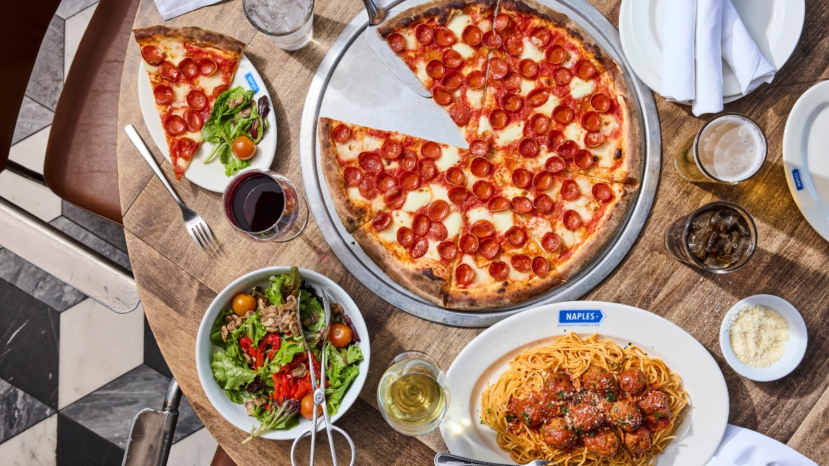 A circular wooden dining tabled is filled with an Italian meal from Naples Ristorante. A huge pepperoni pizza takes up most of the space on the table. Sides include spaghetti and meatballs, a salad filled with vegetables, a small dish of parmesan cheese, and assorted beverages.