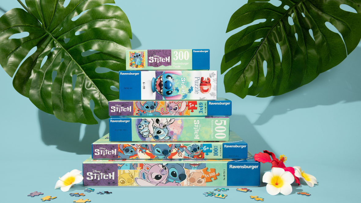 A stack of Lilo and Stitch puzzles from Ravensburger in front of a pale blue background. Two large green leaves frame the stack of puzzles, in the top left and right corners, with a few small colorful flowers and puzzle pieces on the ground in front of the stack.