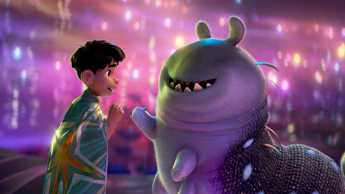 On the left, Elio, a young boy with dark hair wearing a shimmering, star-patterned teal cape, looks up with a hopeful smile. He faces Glordon, a large, round, purple alien with a wide, toothy grin and small antennae. The background is a dreamy, out-of-focus expanse of glowing purple and pink lights.