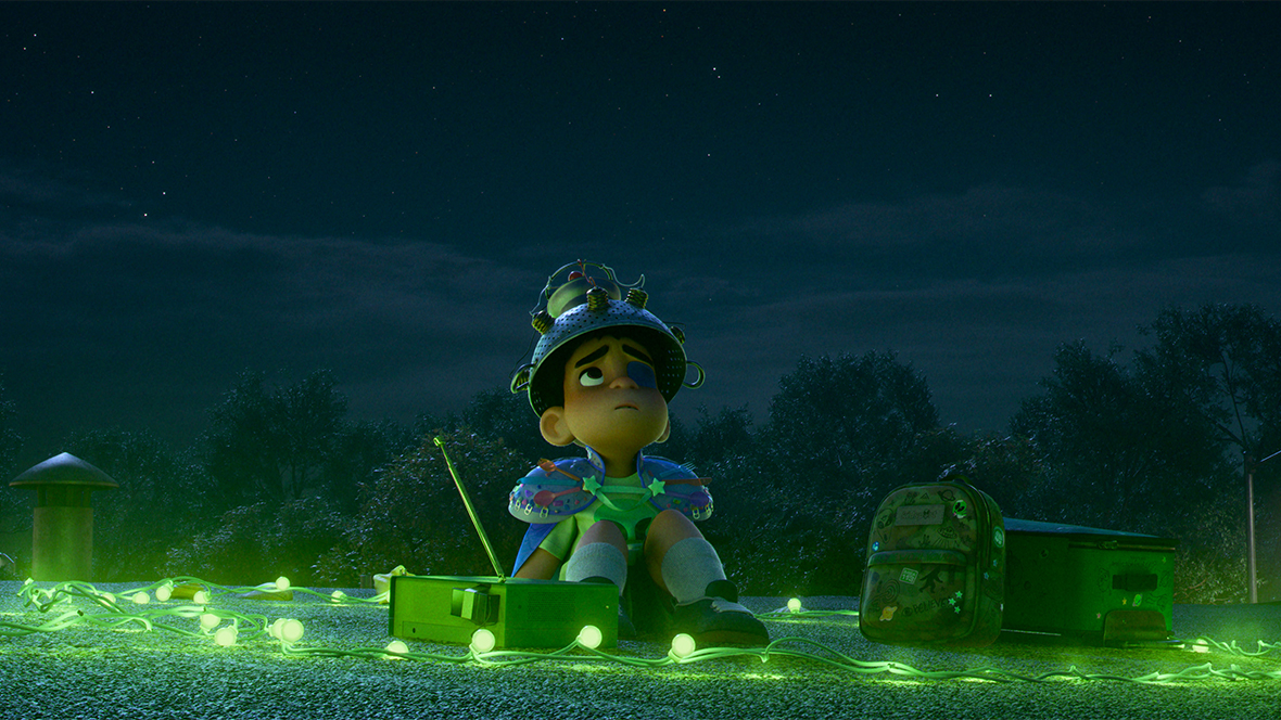 Elio, a young boy with an eye patch, sits alone on a gravel rooftop under a vast, dark, starry sky. He wears a makeshift space helmet made from a metal colander and a blue cape adorned with colorful stickers. Surrounding him on the roof are a tangled string of glowing green lights, a green portable radio with its antenna extended, and his backpack. He gazes upward with a hopeful yet lonely expression. The background shows the dark silhouettes of trees and a single glowing street lamp, all bathed in a mystical green hue.