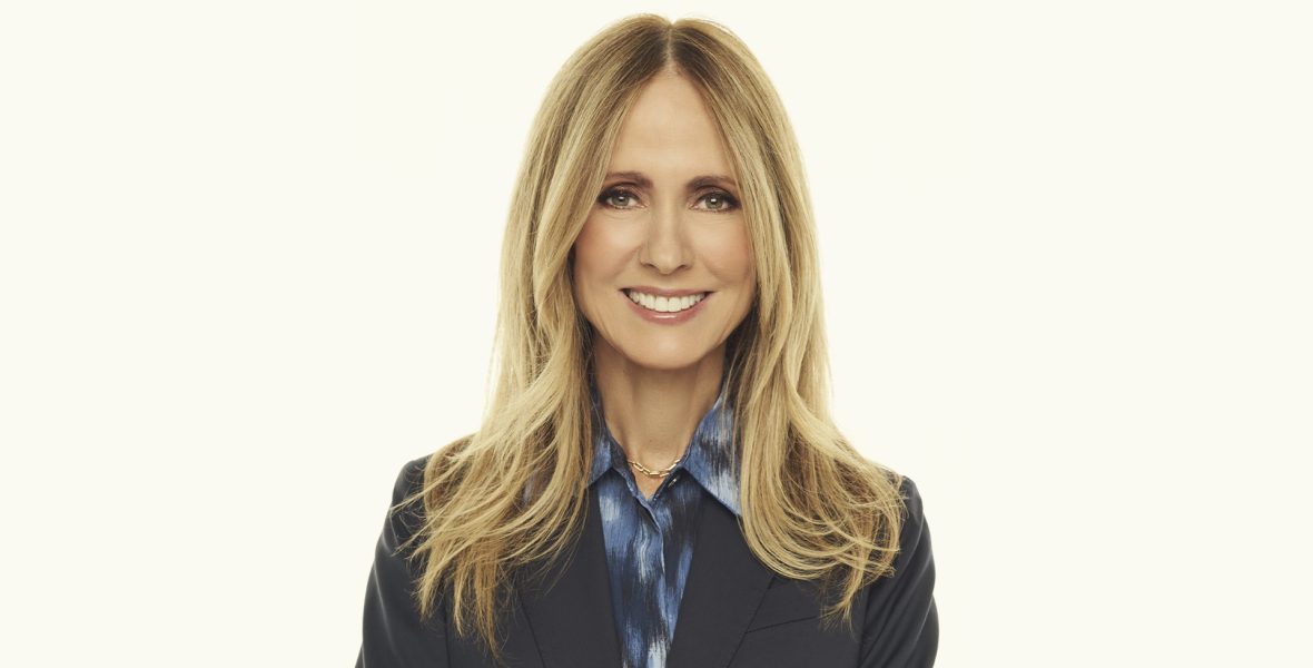 Professional studio portrait of Dana Walden with long blonde hair, wearing a dark blazer over a patterned blue blouse, photographed against a light neutral background.