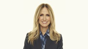 Professional studio portrait of Dana Walden with long blonde hair, wearing a dark blazer over a patterned blue blouse, photographed against a light neutral background.