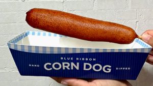 A corn dog from Blue Ribbon Corn Dog floats above Blue Ribbon Corn Dog plating.