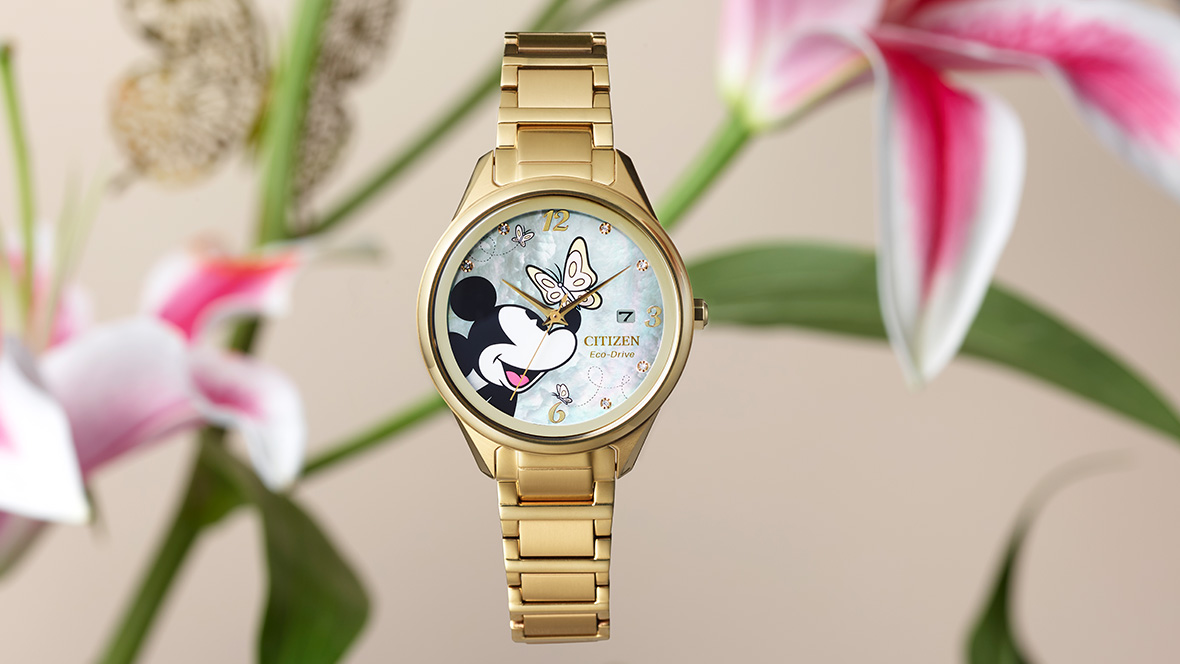 A Citizen Mickey Flutterby watch sits front and center on a background featuring lilies. The watch dial includes a photo of Mickey Mouse smiling, with a butterfly resting on his nose. The color of the dial is a rich pearl color. The watch includes a date window and 12-, 3-, and 6-hour markers. The case and bracelet are a gold color.
