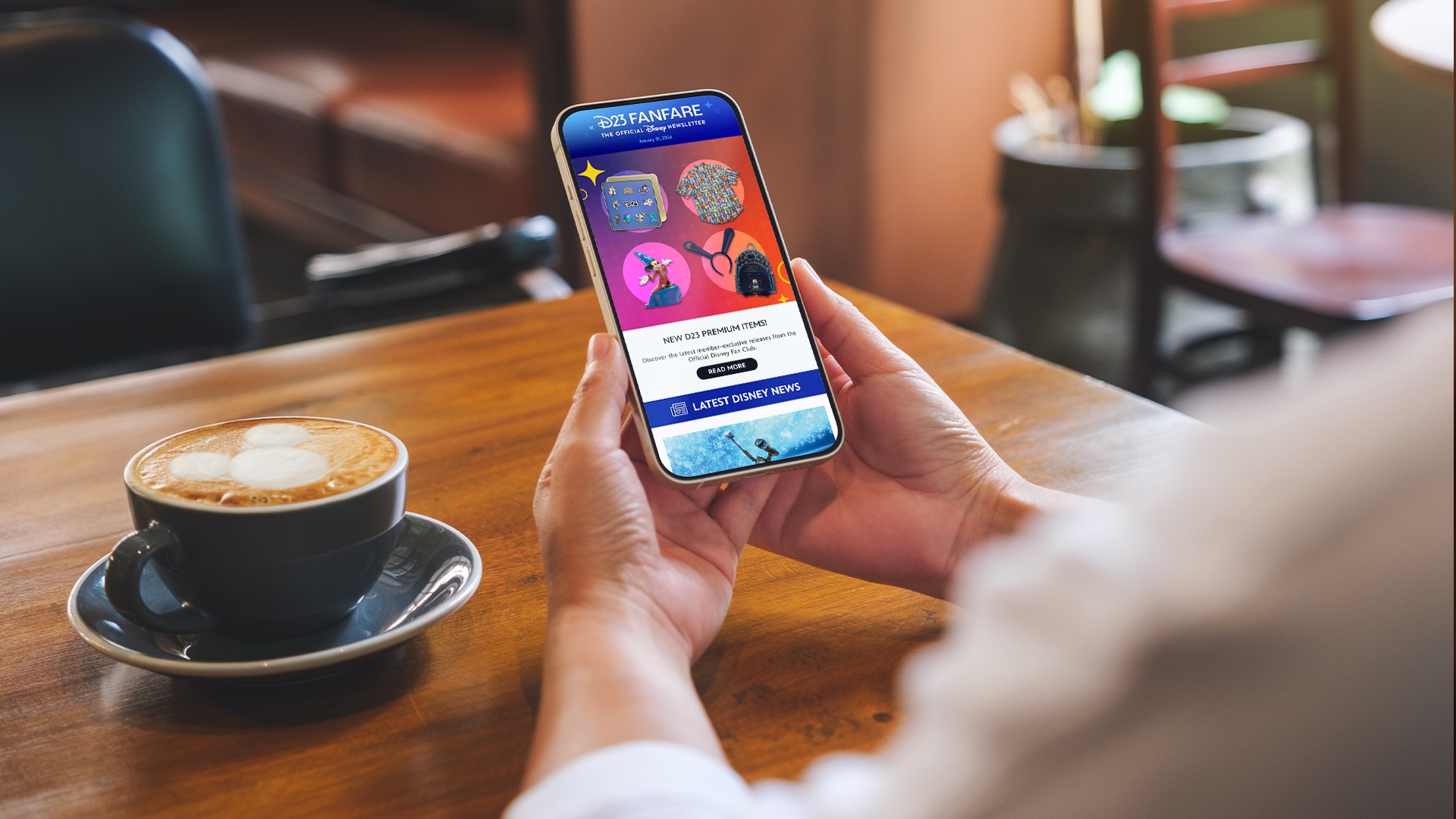 Person holding a smartphone at a caf&eacute; table, viewing the D23 Fanfare newsletter on screen with featured D23 merchandise and Disney news, next to a cup of coffee.