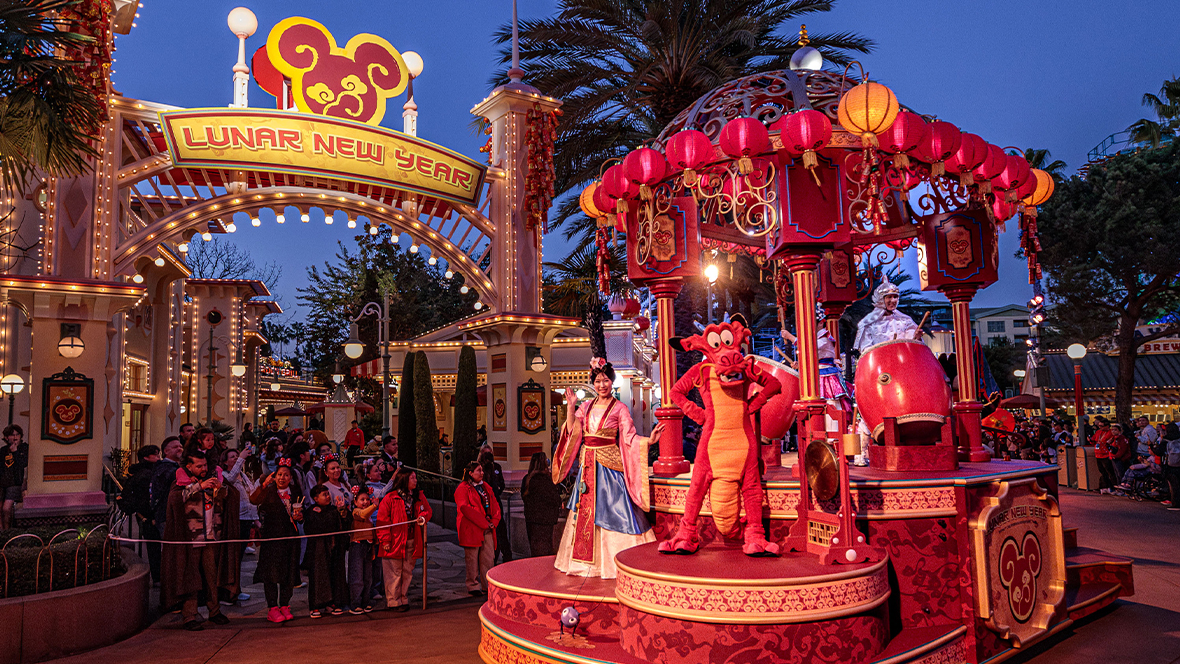 An image of Mulan&rsquo;s Lunar New Year Procession at Disney California Adventure Park during the limited-time Lunar New Year celebration. A crowd of guests to the left of the image, under a Lunar New Year sign, is watching and taking photos of the procession, which is coming into frame on the right.
