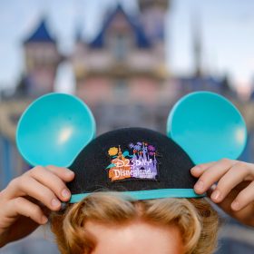 A person adjusts blue D23 Day at Disneyland Resort Mickey ears in front of Sleeping Beauty Castle during D23 Day at Disneyland.