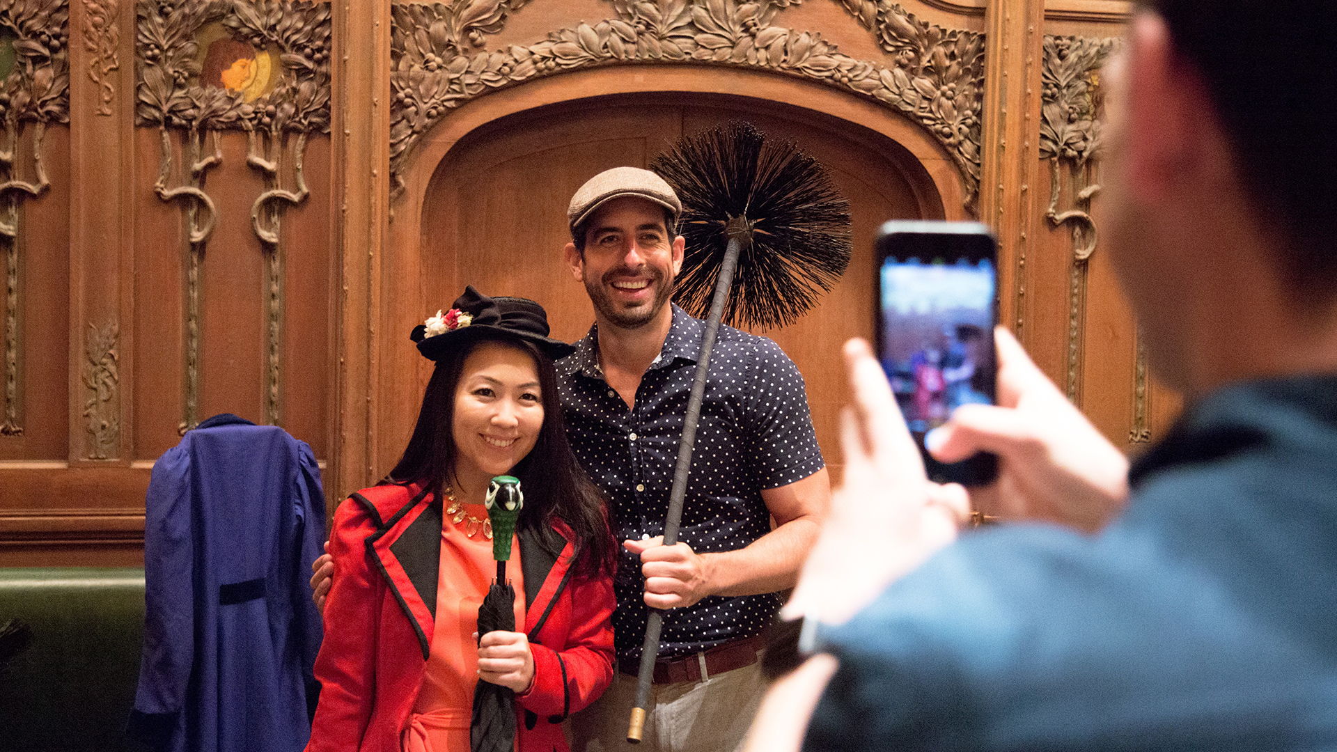 Two people dressed as Mary Poppins and Bert pose with a parrot umbrella and chimney sweep while a guest photographs them during a D23 New Amsterdam tour.