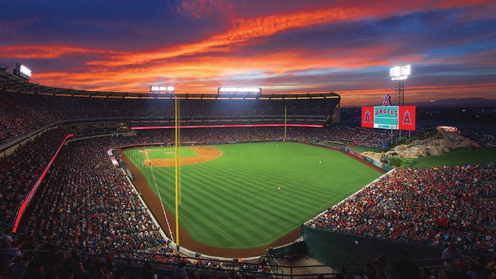 Fans watch a baseball game at Angel Stadium as the scoreboard displays the Los Angeles Angels logo and name at sunset.