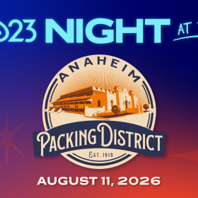 Promotional graphic for D23 Night at the Anaheim Packing District. The top of the image features the D23 logo and the words “Night at the” in blue lettering; centered on the image is the Anaheim Packing District emblem showing an illustrated historic building with the text “Anaheim Packing District, Est. 1919.” At the bottom, the event date “August 11, 2026” appears in white. Everything is set against a gradient background of deep blue to orange-red.