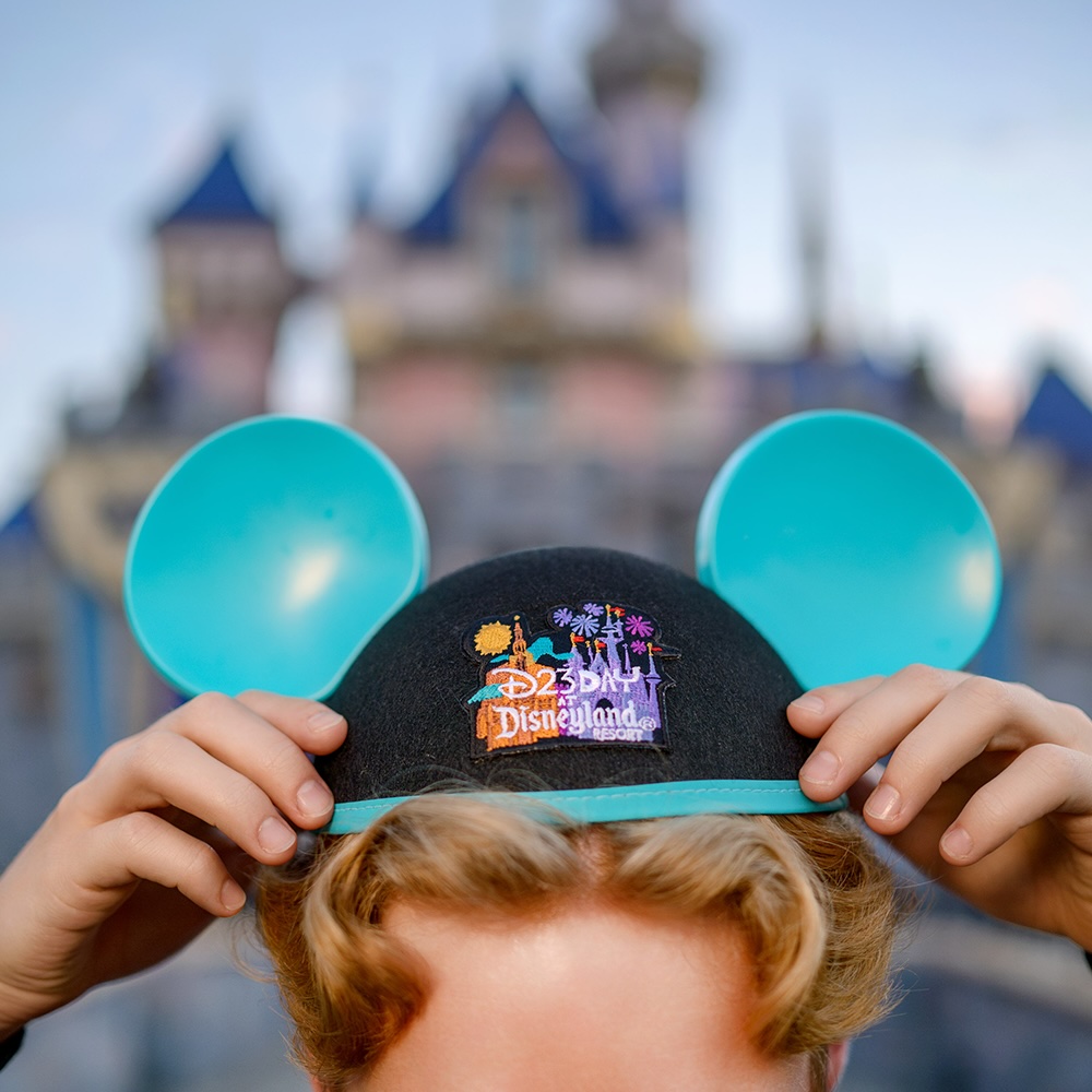 A person adjusts blue D23 Day at Disneyland Resort Mickey ears in front of Sleeping Beauty Castle during D23 Day at Disneyland.