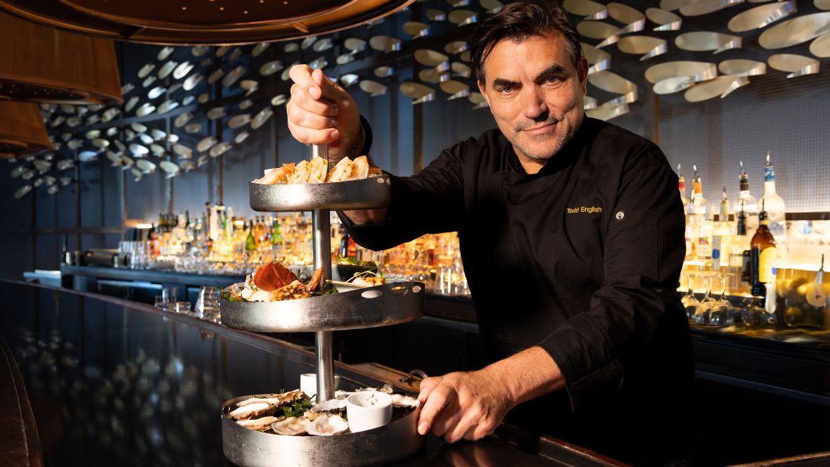 Chef Todd English presenting a multi-tiered seafood tower at a modern bar, with bottles and warm ambient lighting in the background.