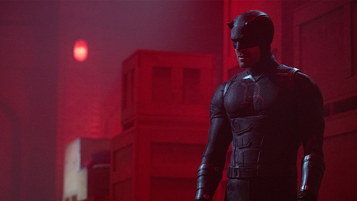 An image from Daredevil: Born Again season 2, featuring Charlie Cox as Daredevil. He is in his Daredevil suit and standing in front of stacked boxes.