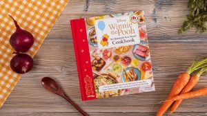 An image of the cover of Winnie the Pooh: The Hundred-Acre Wood Cookbook by James Asmus, Liz Tarpy, and Vivian Jao. Towards the top of the cover is the logo of the book&rsquo;s title, alongside an image of Pooh tied to a blue balloon. The majority of the cover features many dishes seen inside the cookbook, displayed on a large table. The cookbook itself is sitting on a wooden table, surrounded by a ladle and some carrots and onions.