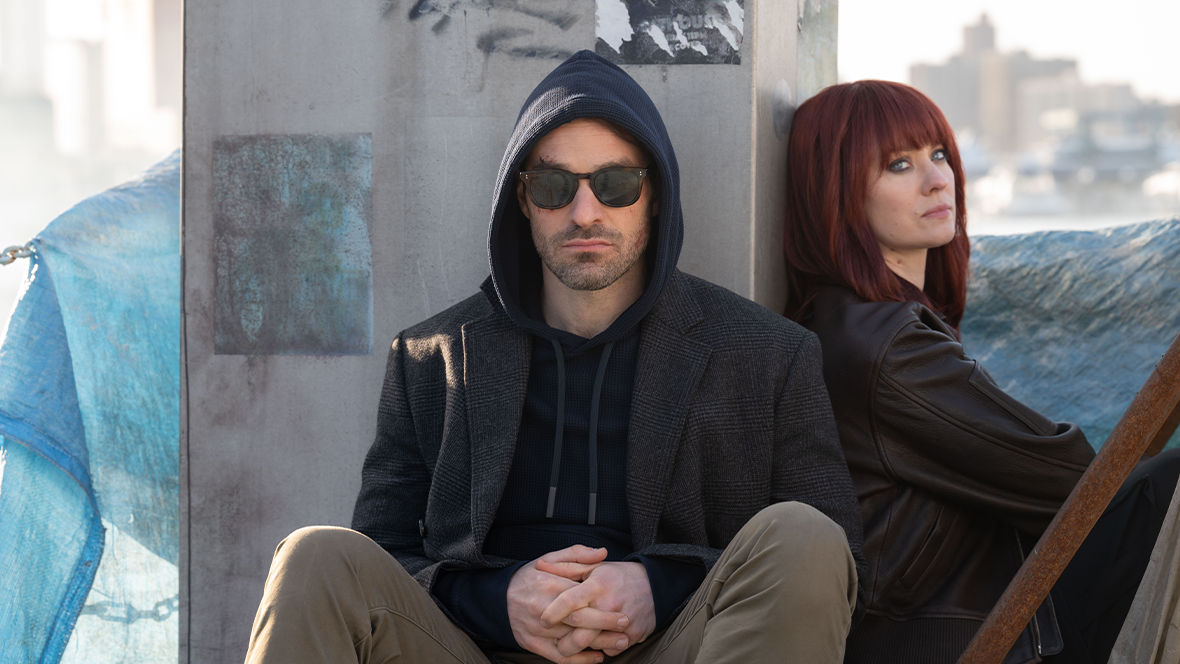 An image from Daredevil: Born Again season 2, featuring Charlie Cox as Matt Murdock (left) and Deborah Ann Woll as Karen Page (right). They are sitting against a column of a building and looking towards the camera.