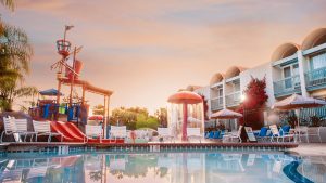 The pool of the Howard Johnson Anaheim Hotel sits next to the hotel rooms, just as the sun is setting. The pool features a pirate-themed water playground, including a crow&rsquo;s nest and a slide. Scattered throughout the pool areas are tables, chairs, and lounge chairs.