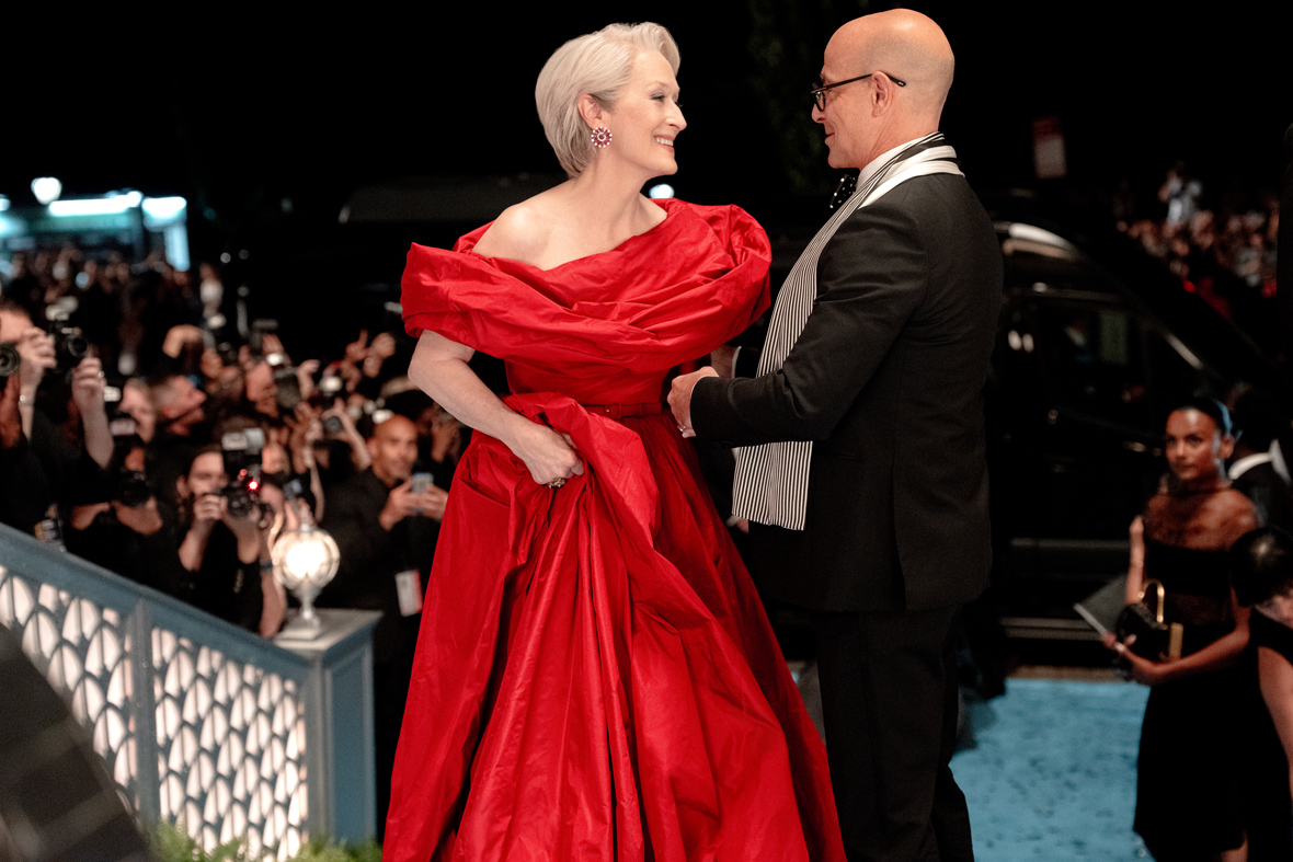 An image of Meryl Streep as Miranda Priestly, in a red couture ball gown, and Stanley Tucci as Nigel, in a tuxedo, from The Devil Wears Prada 2; they are standing on a red carpet, and paparazzi are seen in the background.