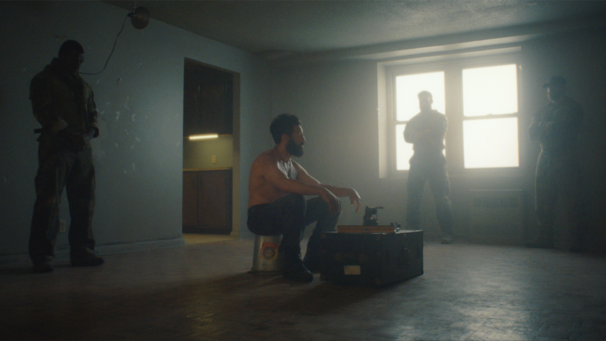 Frank Castle/The Punisher, played by Jon Bernthal, is shirtless and seated on a bucket in a scene from A Marvel Television Special Presentation: The Punisher: One Last Kill on Disney+. The room is nearly empty, and he is surrounded by three former Marines.